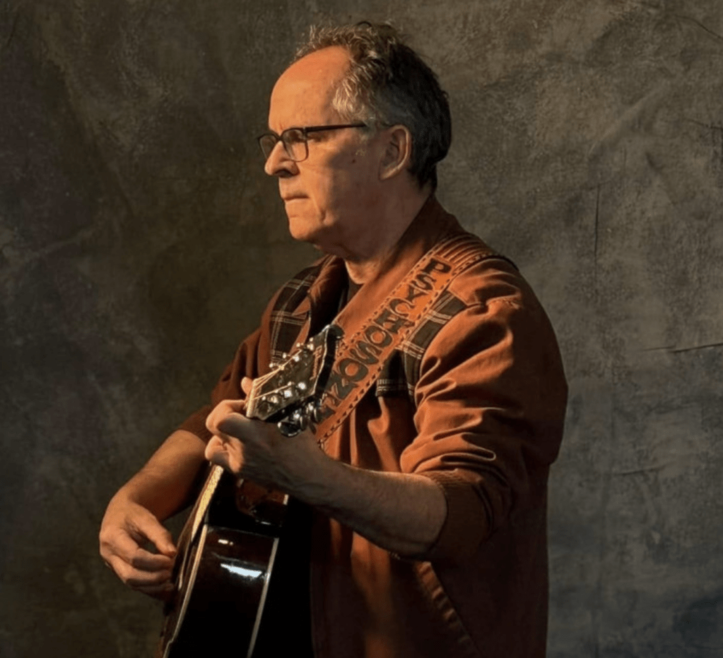 A standing man in a red long sleeve shirt facing to the left and playing acoustic guitar.