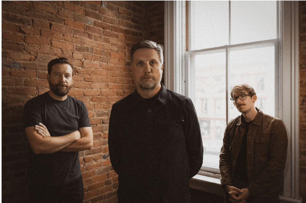 Three men in dark clothing, standing in front of a backlit window and brick wall, looking at the camera 