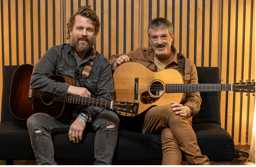 Two men sitting on a couch with acoustic guitars - they are smiling and dressed in jeans and jackets 