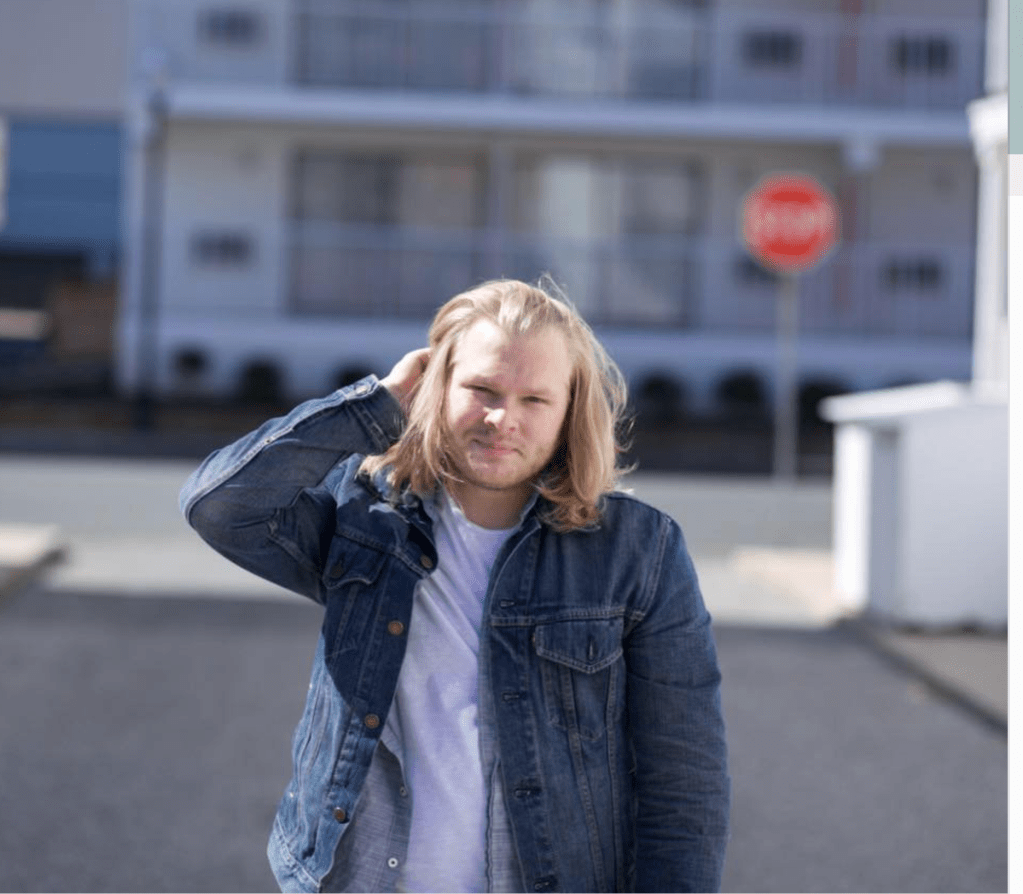 A blond man dressed in a jean jacket and while t-shirt looks at the camera.  