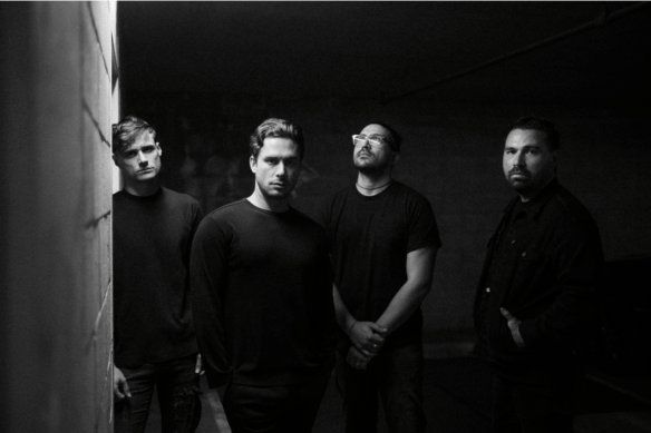 Four men standing in a dimly lit area. They are all dresses in black.