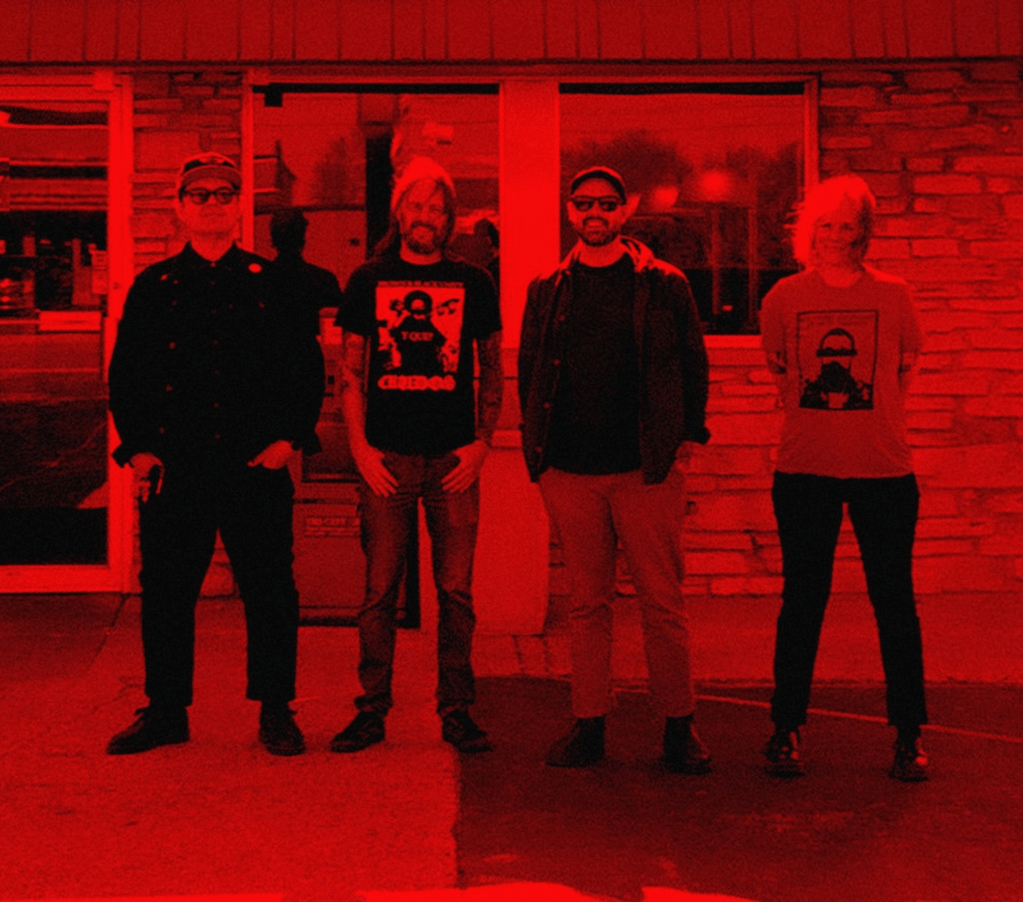 Three men and one woman stand outside a shop that looks like a convenience store.  They look happy.  