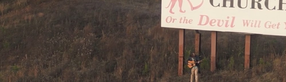 A man, holding a guitar on a hillside. He is standing underneath, and looking up at, a large billboard.