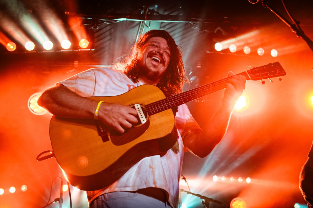 A male musician on a brightly lit stage, smiling broadly and playing guitar.