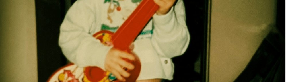 A young boy stands clutching a red and white toy guitar. He is grinning widely and looks happy.