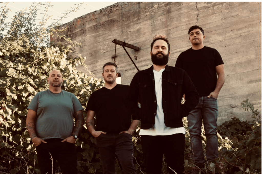 Four men, casually dressed and standing in front of an old concrete wall, look out toward the viewer.