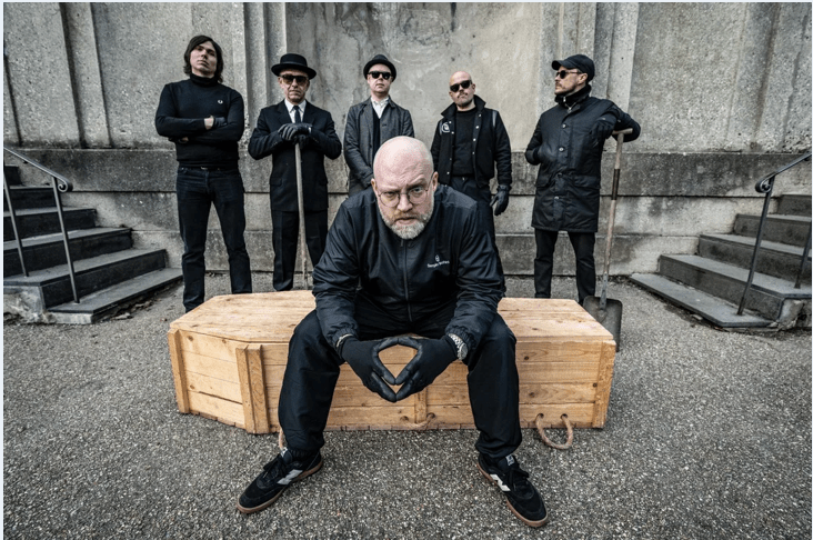 Six men, one sitting on a wooden casket and the other five standing a few feet behind, look out toward the viewer.  They are all dressed in black and the mood is kind of somber.  