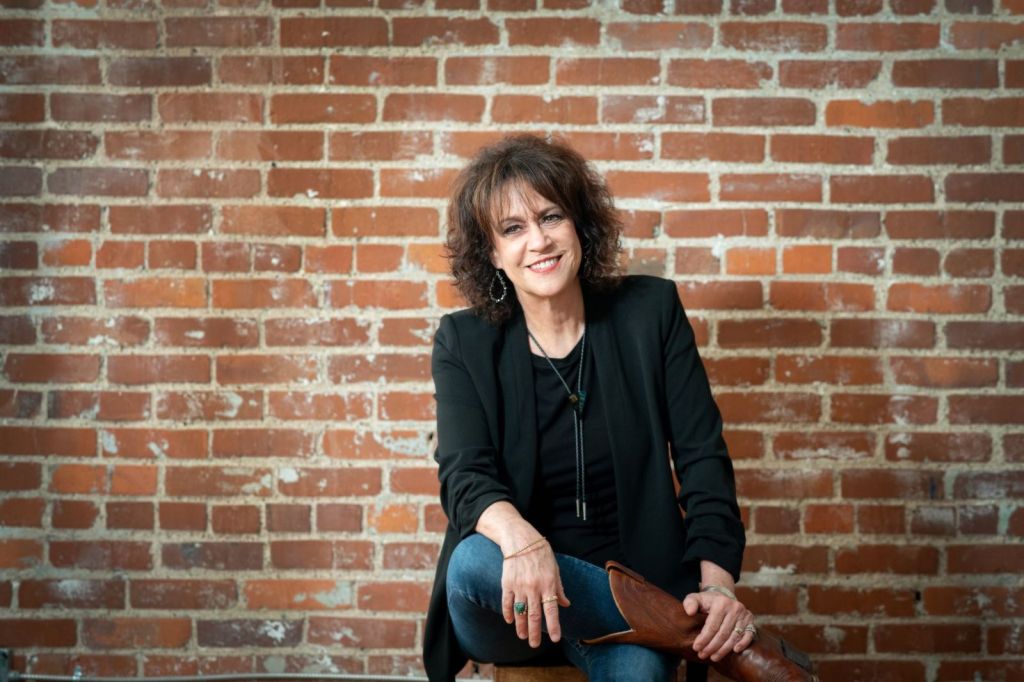 A woman, dressed in a black jacket, jeans and tall boots, sits against a red brick wall. She is smiling and looking slightly off to the viewers left.