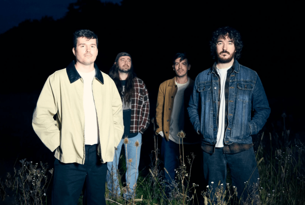 Four casually dressed men, front lit as the scene is dark, stand in a field looking forward the viewer.