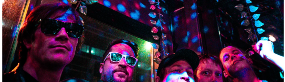 Five men, standing slightly off-center from camera in a disco lit environment. They look slightly amused.