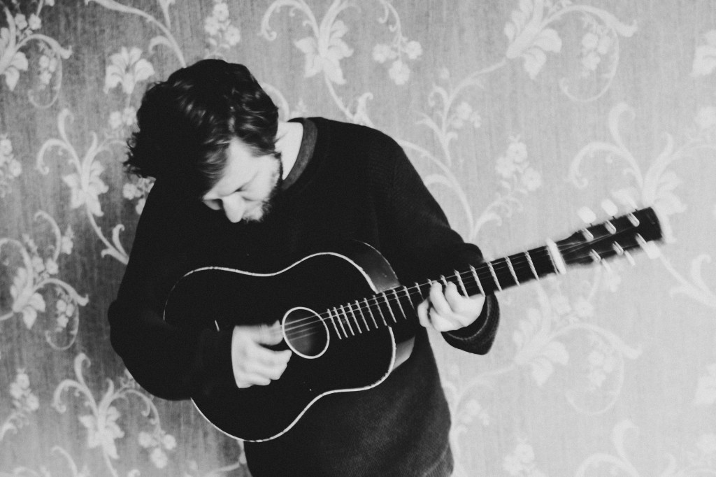 A black and white image of a man standing against a floral print wall.  He is dressed casually and playing an acoustic guitar.  