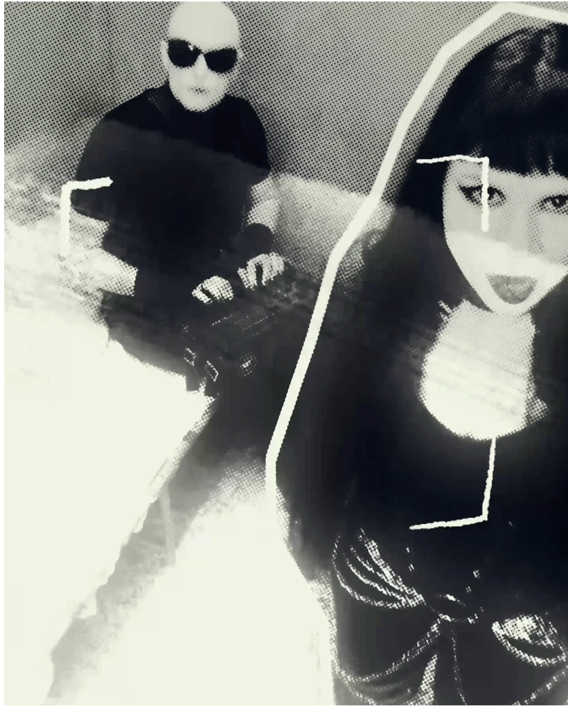 Two people (one sitting on a cot and one standing) inside a prison cell, both of them looking toward the viewer. The person on the bed has a musical device on their lap.