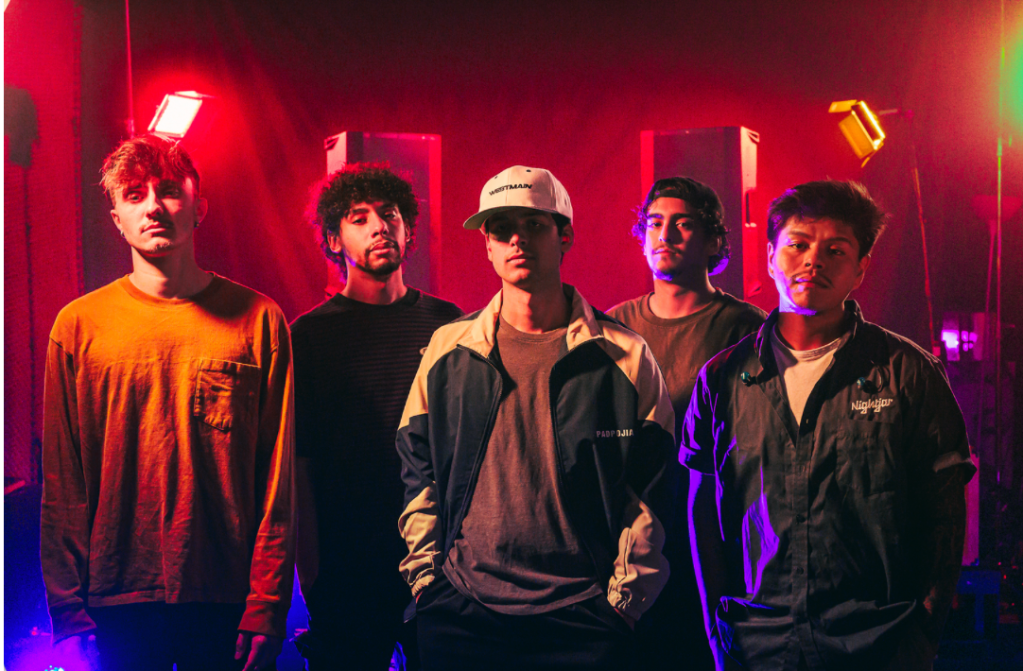 Five young men, casually dressed and standing in front of a music stage, look out toward the viewer.  