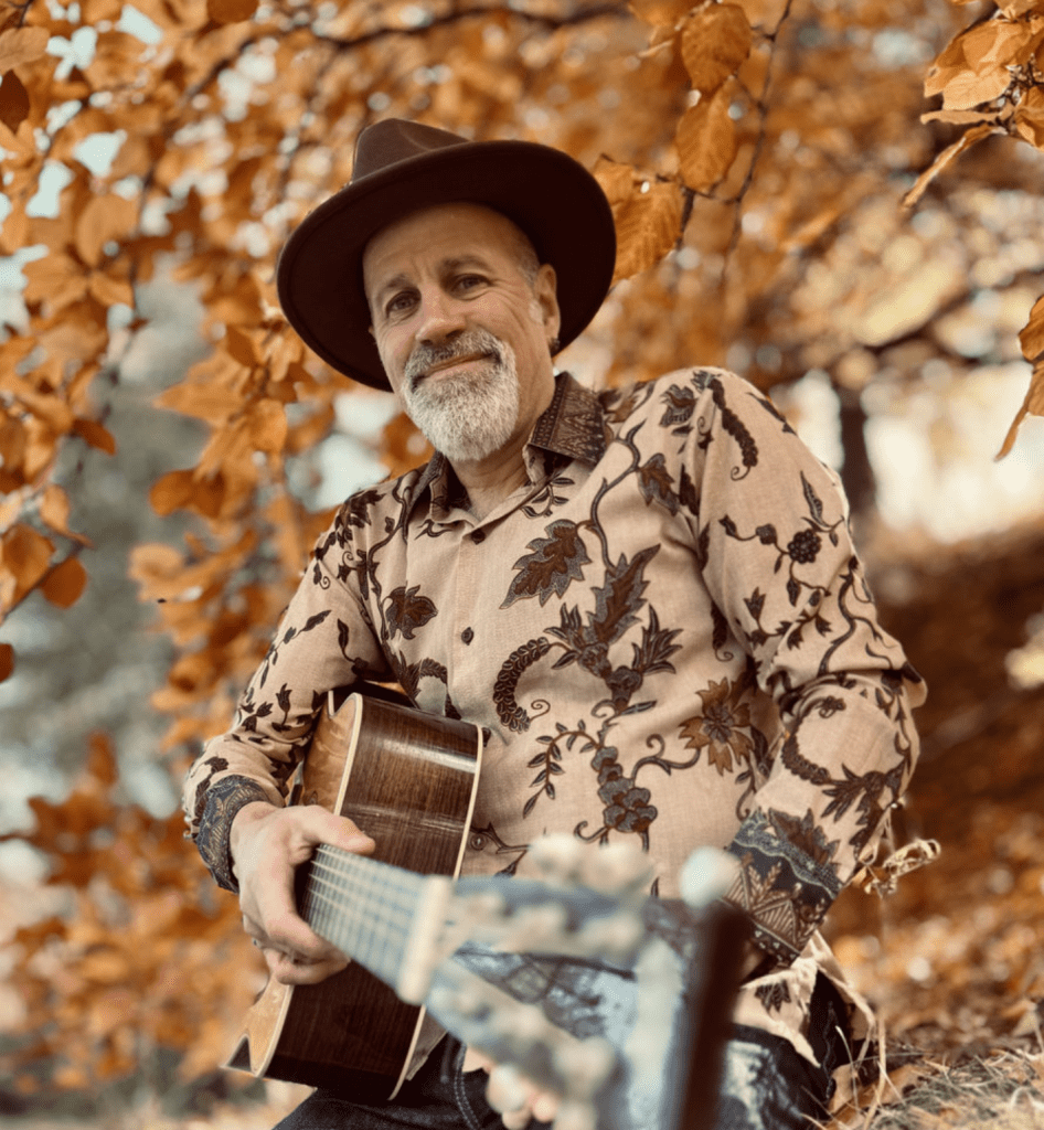 A man dressed in a patterned, long sleeve shirt and famed by an autumn tree looks toward the viewer.  He is holding an acoustic guitar.  