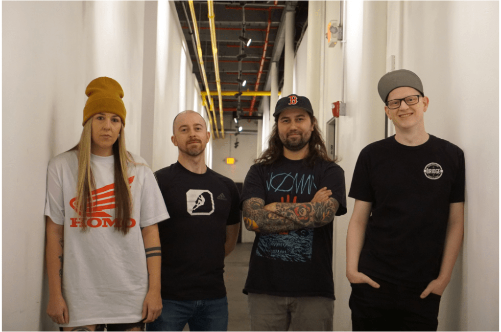 One woman and three men, all casually dressed, stand in a long hallway looking out toward the viewer.