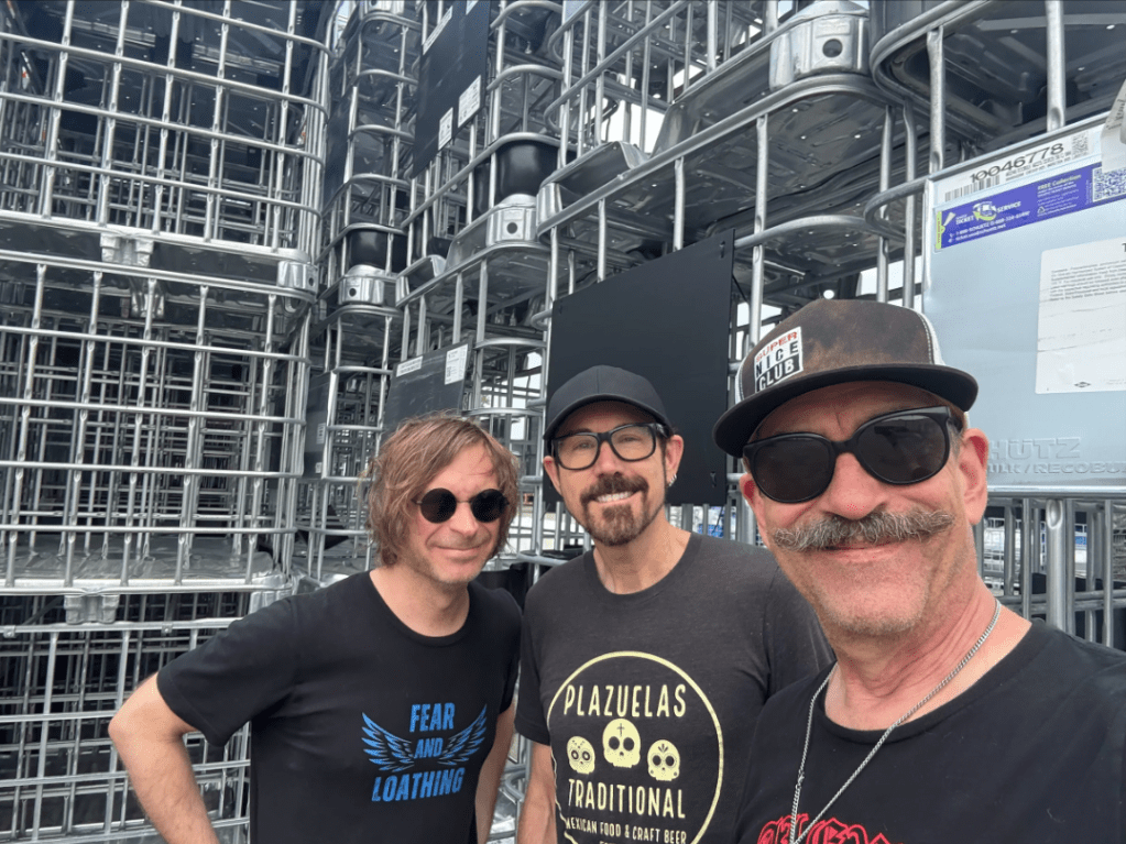 Three men dressed in black t-shirts (with lettering) look out at the viewer.  They appear to be pretty happy.  