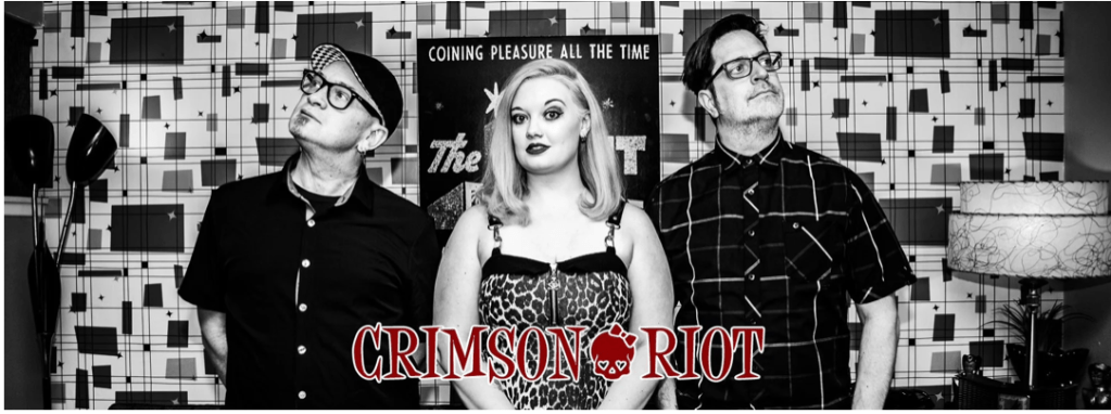 One woman and two men stand against some a wall with busy wallpaper.  The woman is looking at the viewer while the men look up and off to the left and right.  