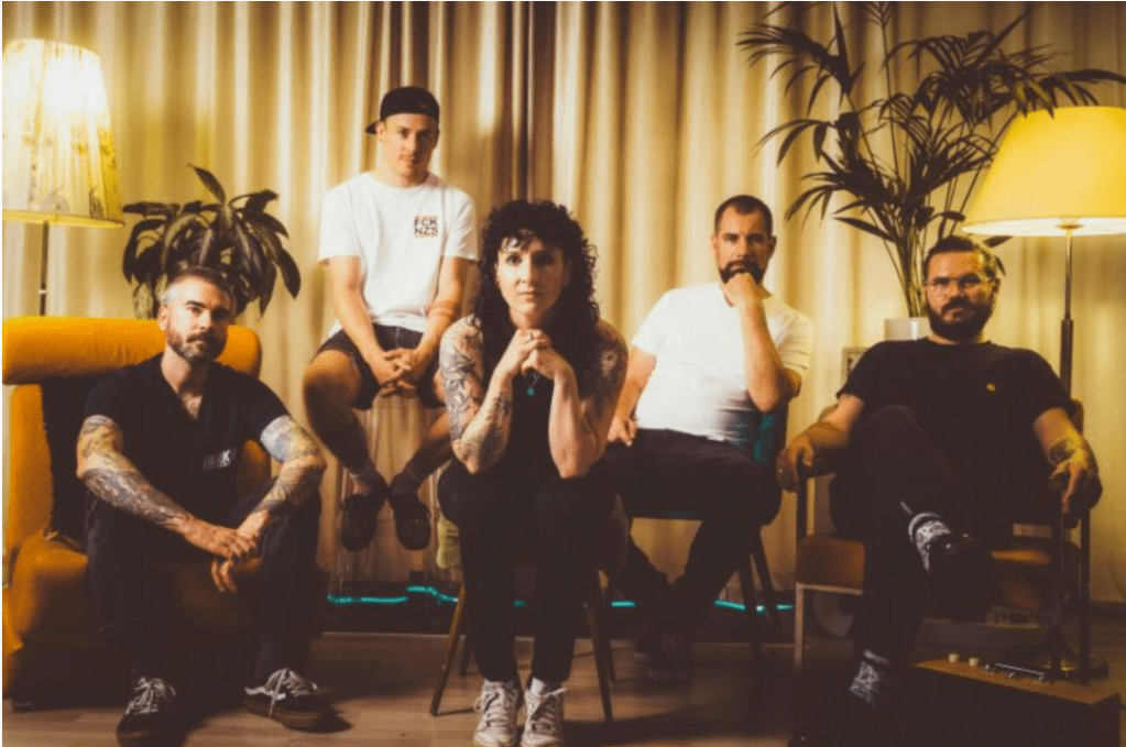 Four men and one woman sit in what looks like an older style living room setting.  They are looking out toward the viewer with neutral expressions.  