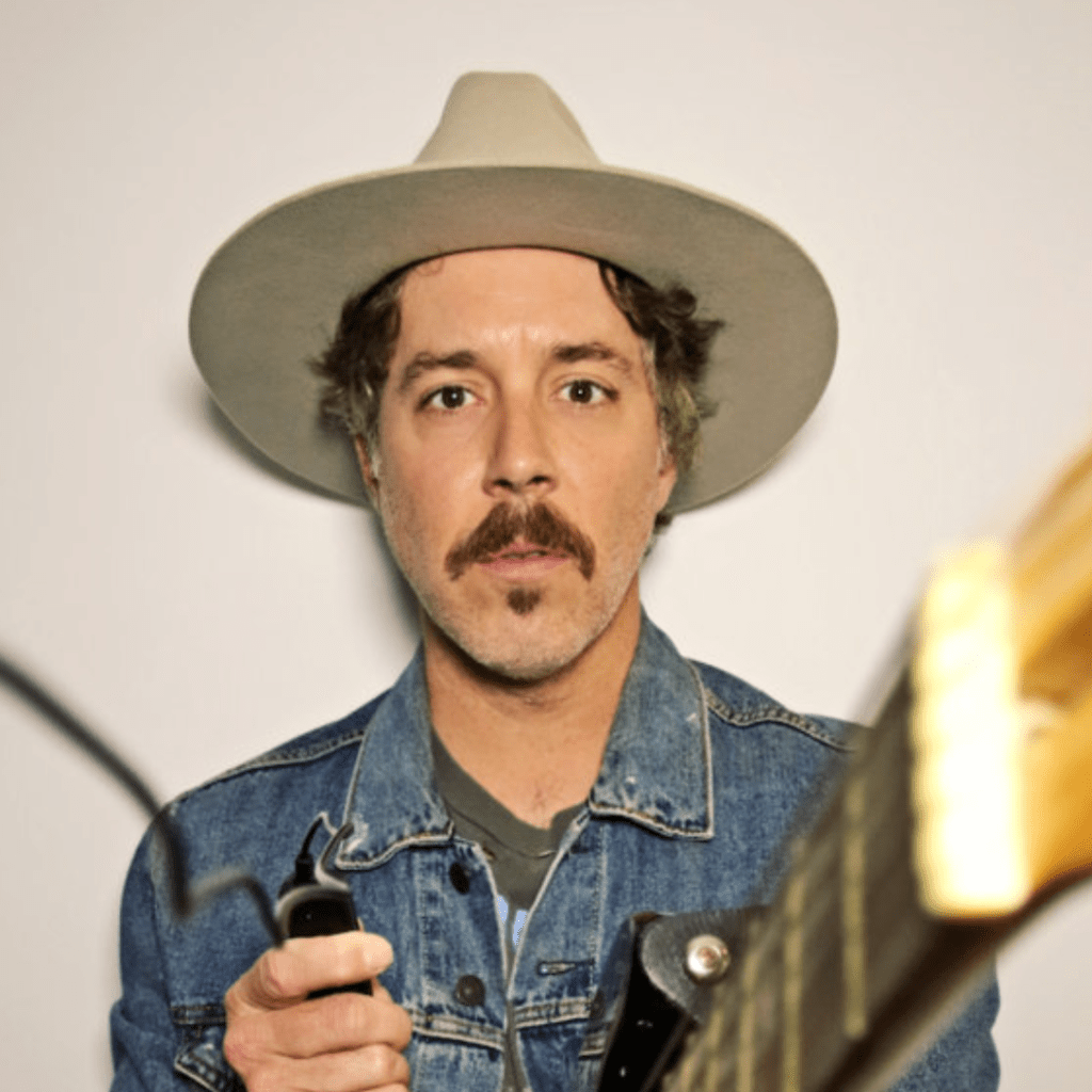 A man wearing a hat and denim jacket looks out toward the viewer.  He is also holding a guitar that is pointed toward the camera.