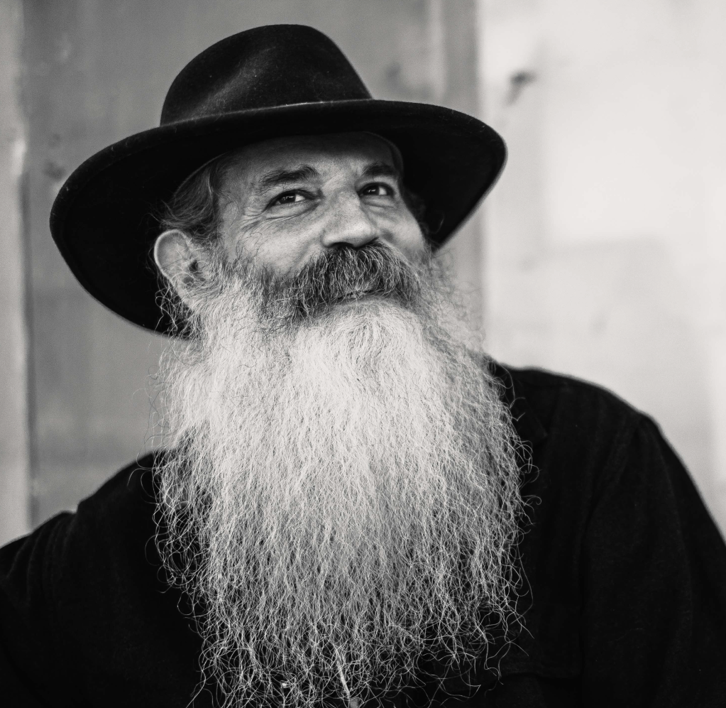 A beareded man in a hat looks off to the viewers left. He looks happy.