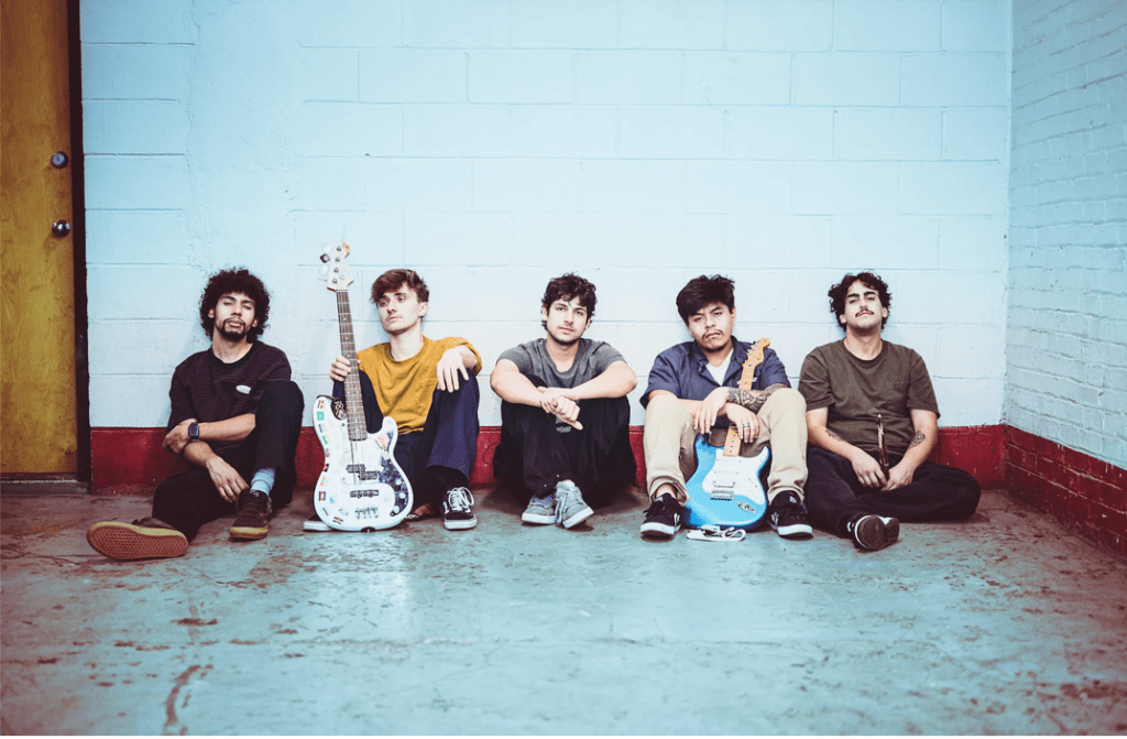 Five young men, casually dressed and sitting against a brick wall. Two of them hold stringed instruments.
