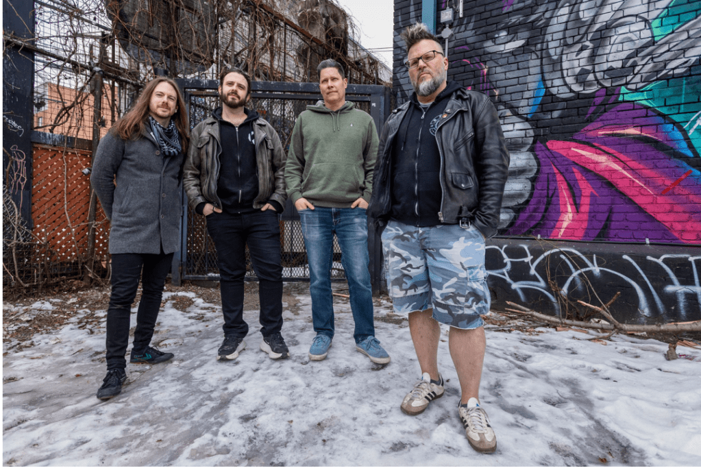 Four casually dressed men stand in front of a tall fence and a wall with colorful graffiti. 