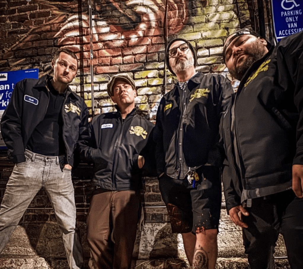 Four men standing against a graffitied brick wall look slightly downward toward the viewer.   They appear a little defiant and edgy. 