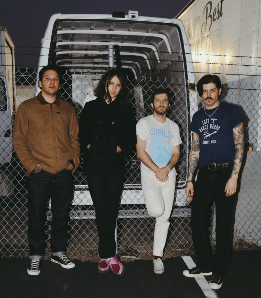 Four casually dressed men stand in front of a barbed wire fence and are looking toward the viewer.   On the other side of the fence is a backed in van with the rear door open.  