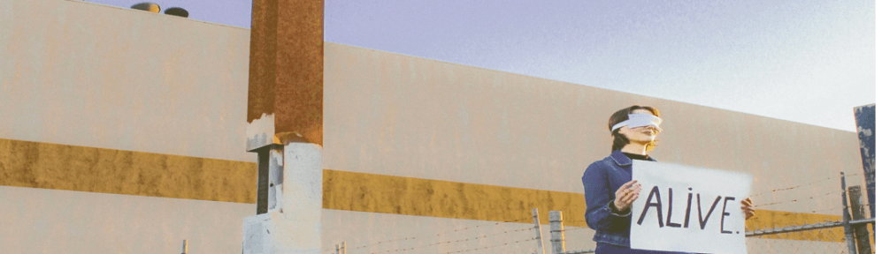 A blindfolded woman sits on top of an electrical power box outside an old motel with a tall sign.