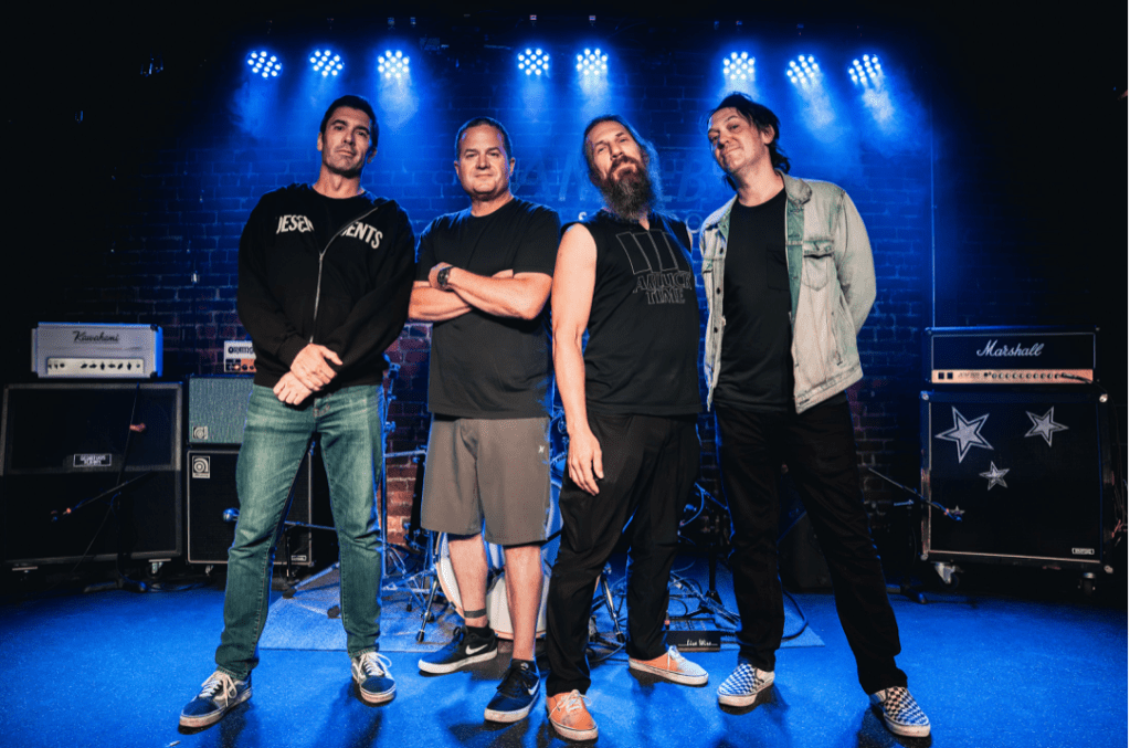 Four men stand on a music stage backlit by the spotlights.