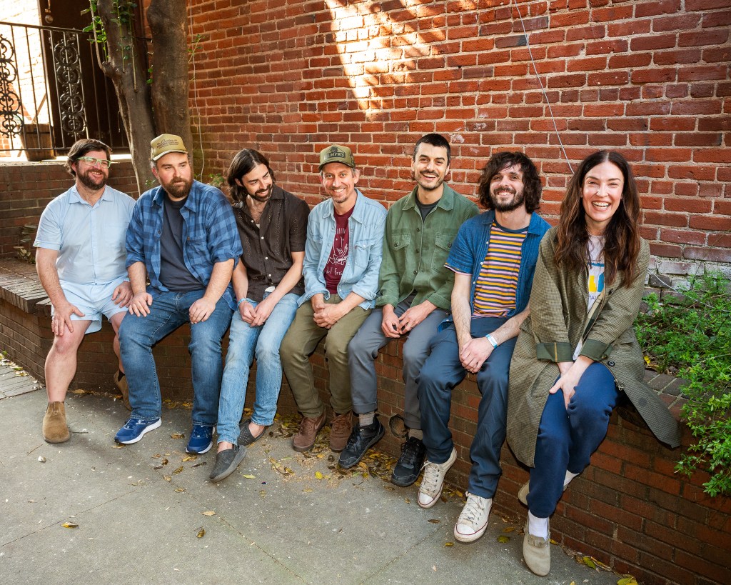 One woman and six men are seated on a low brick bench in front of a similar wall.  They all appear happy.  