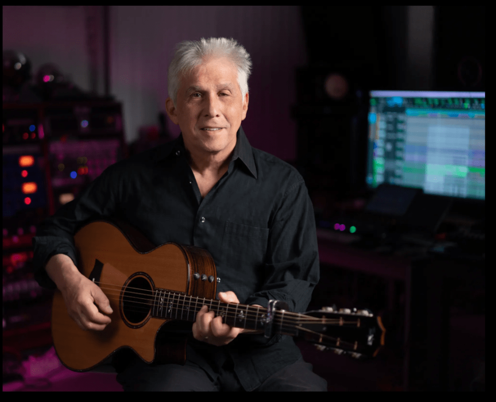 A man sits surrounded by studio recording equipment. He is holding an acoustic guitar.