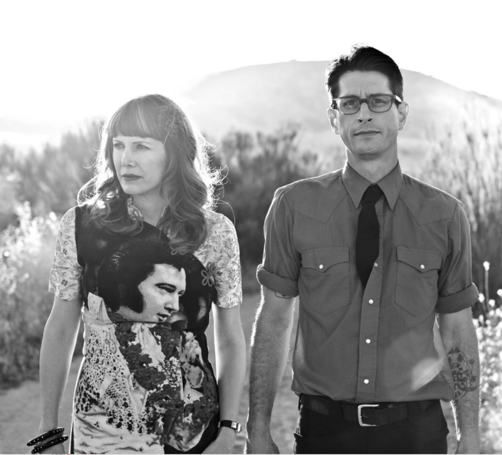 A black and white photo of a man and a woman standing in the near foreground of the image.  Behind them there are scattered bushes along with a hill in the background.