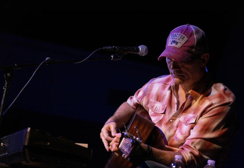 A man in a baseball cap is seated and playing an acoustic guitar.