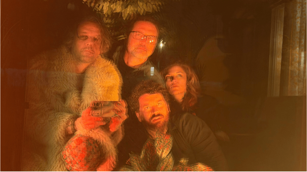 An out of focus color image of four people (three men and one woman) front lit by an orange light and looking toward the viewer.  