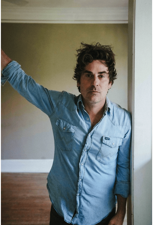 A man in a light blue button down, collared shirt standing in an interior doorway. 